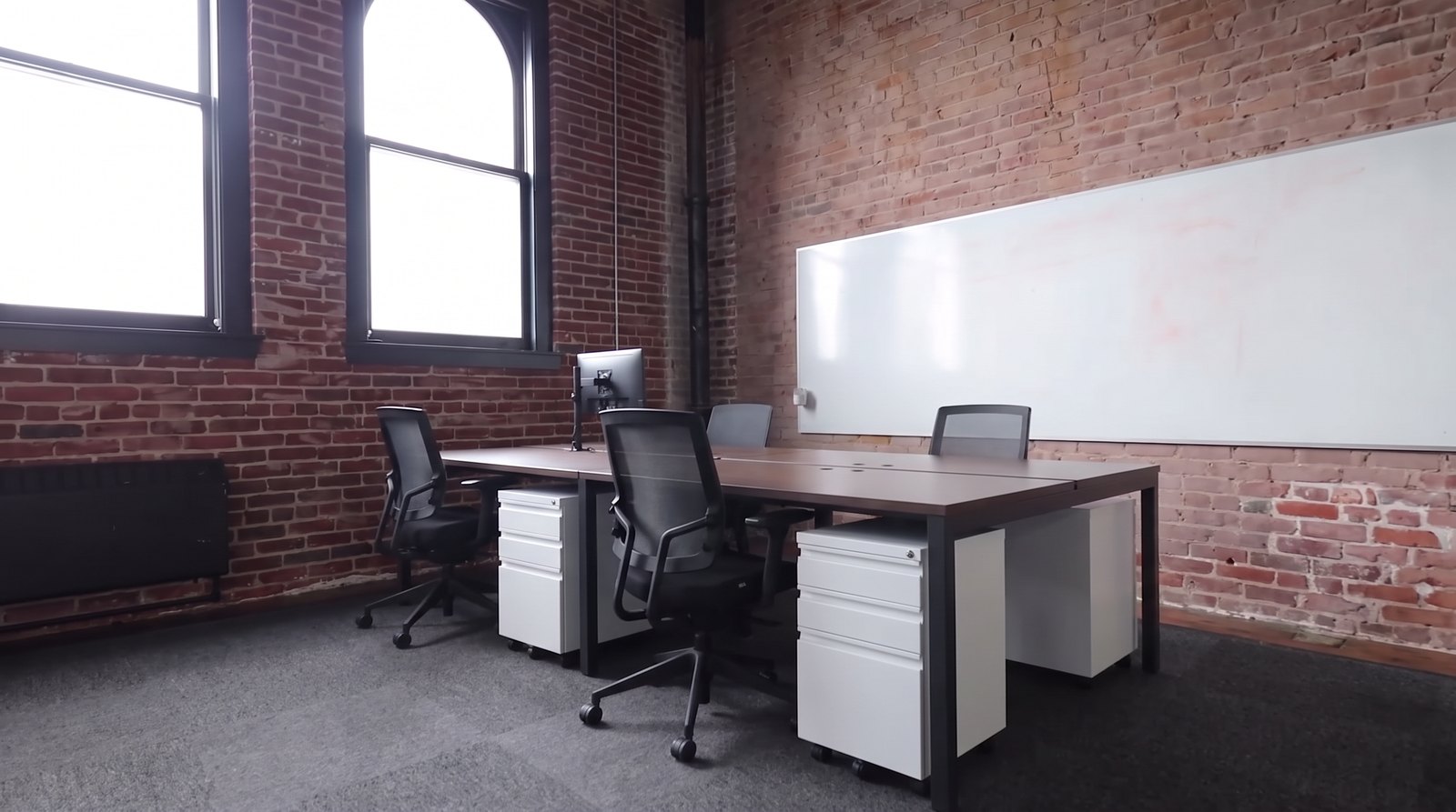 A private office with two arched windows, exposed brick and a whiteboard, shown furnished as an example with walnut topped desks on black frames, white pedestals and black mesh task chairs