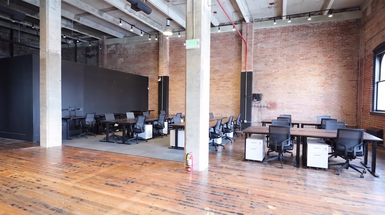 The secondary space shown furnished as an example: rows of walnut topped desks with black frames, white pedestals and black mesh task chairs on the open timber floor between concrete columns